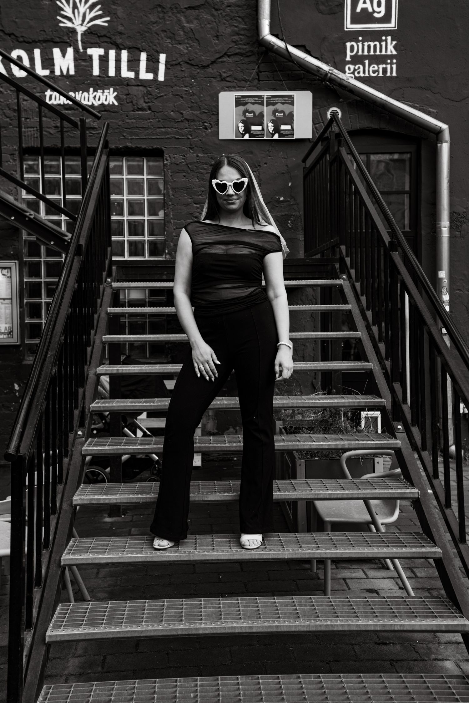 Figure in black outfit and sunglasses poses on metal stairs outside dark building with signage in moody monochrome shot.