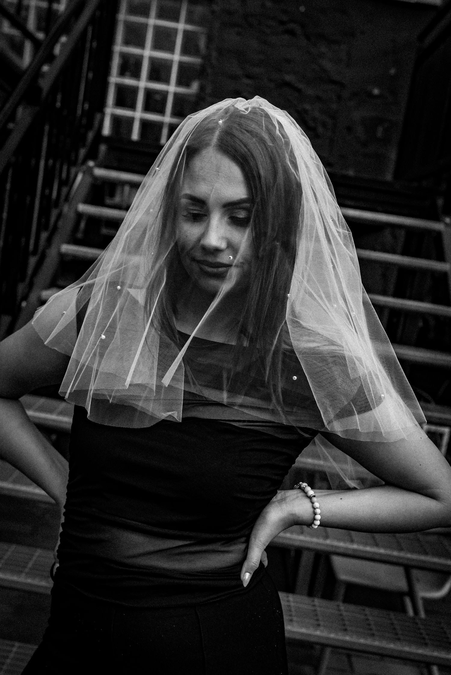 A woman in a black outfit and bridal veil poses on outdoor steps in a moody black and white photograph.