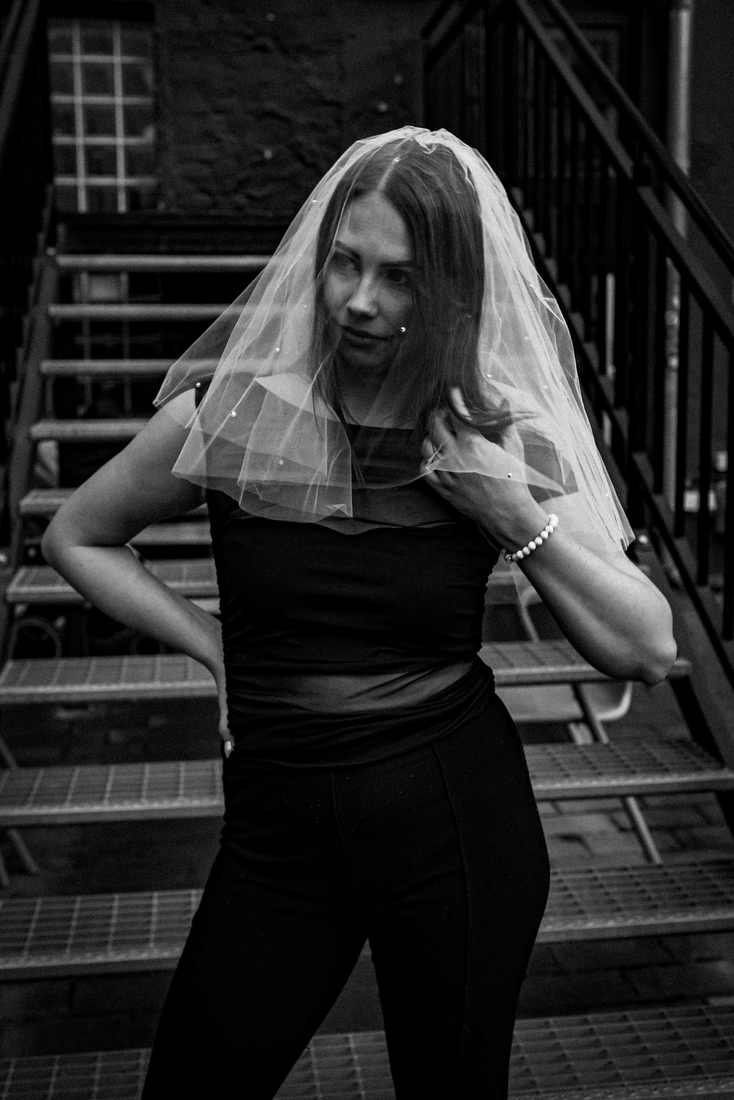 A striking black and white photo of someone in a sheer bridal veil and dark outfit posing on an urban staircase.