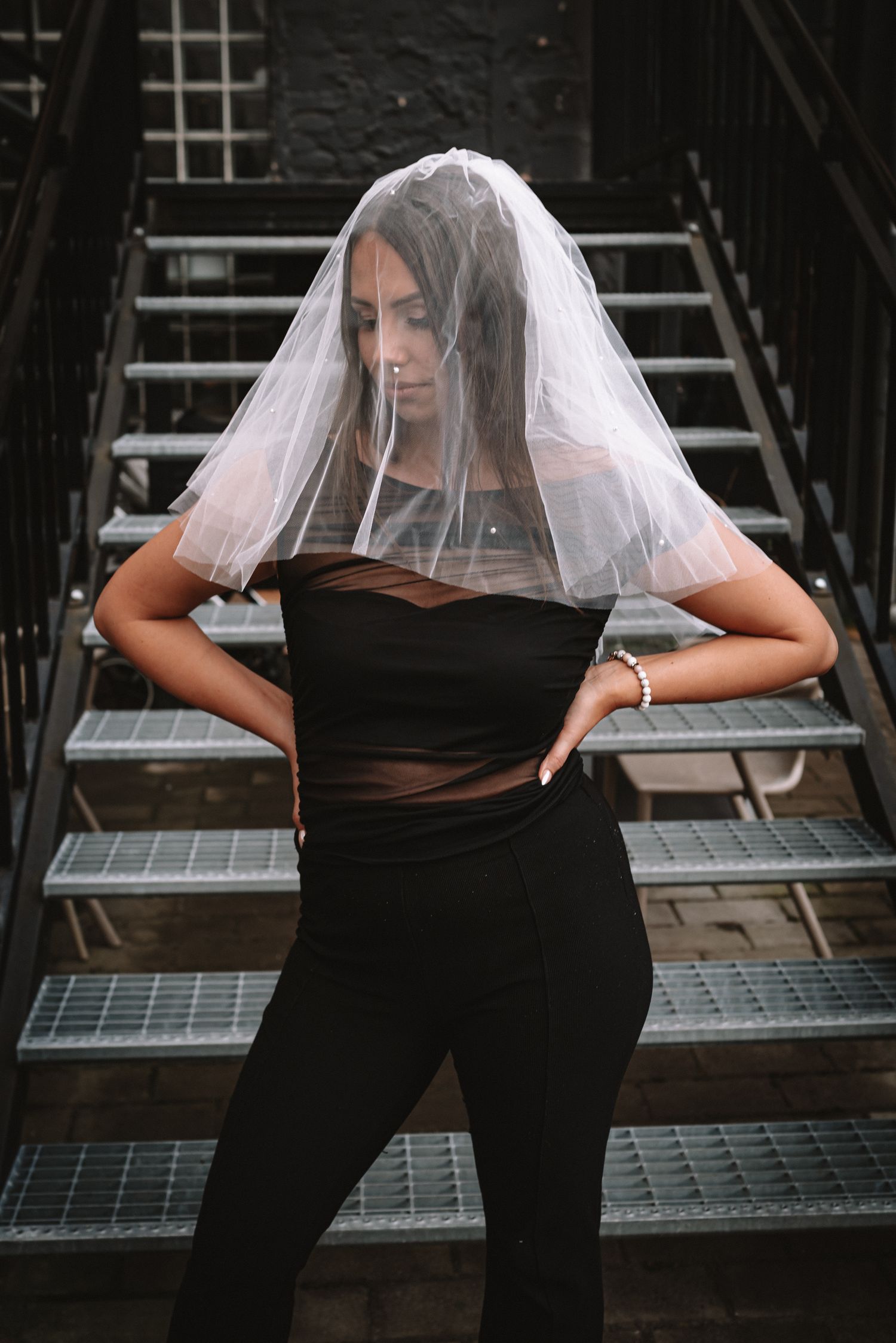 Figure wearing a bridal veil and black outfit poses on an industrial metal staircase in a moody fashion shoot.
