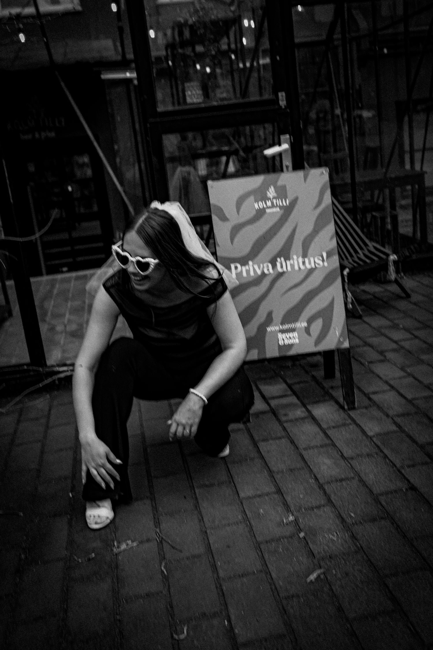 A figure in sunglasses and dark clothing crouches on a cobblestone street next to a promotional sign in monochrome style.