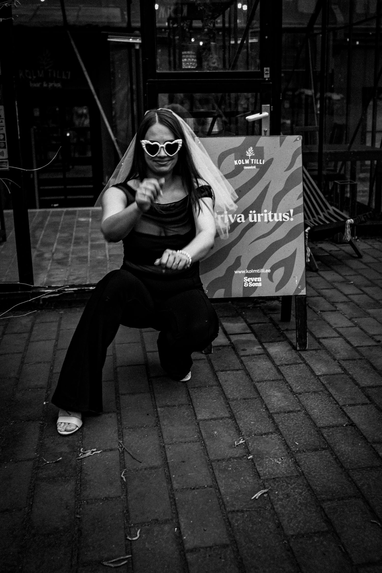 A street fashion pose with sunglasses and black outfit against an urban backdrop with signage in black and white.