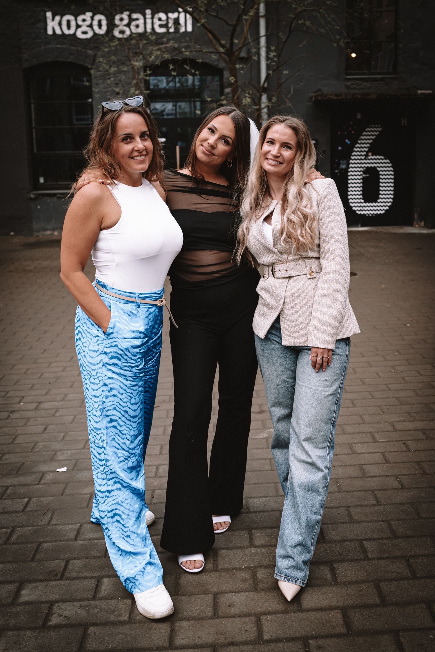 Three friends pose together outside the Kogo Gallery storefront wearing trendy outfits on a cobblestone street.