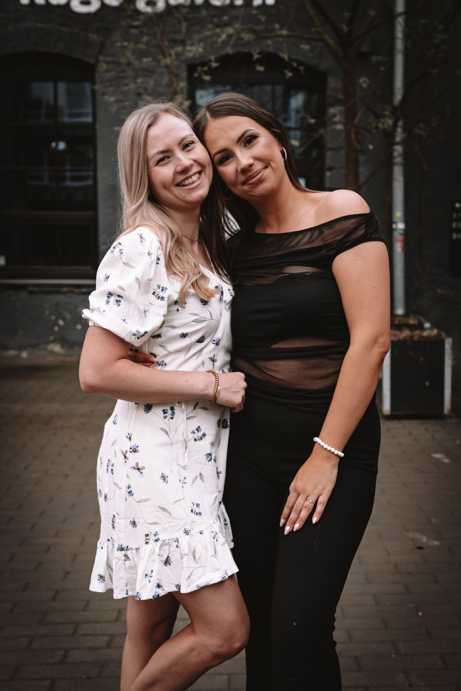 Two friends pose together on a brick walkway, one wearing a white floral dress and one in a black evening gown.