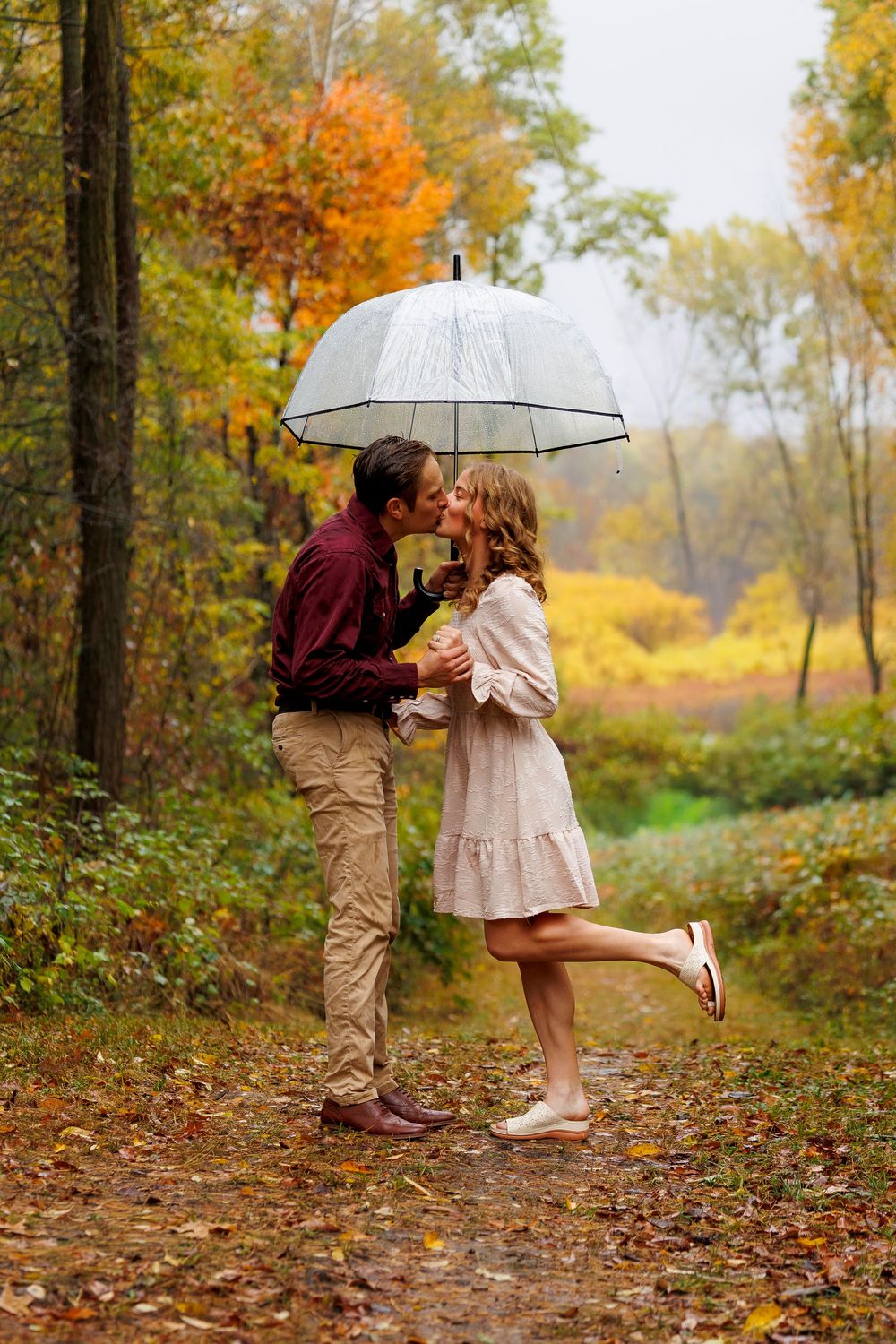 A romantic couple shares tender moments under a clear umbrella on a path covered in autumn leaves.