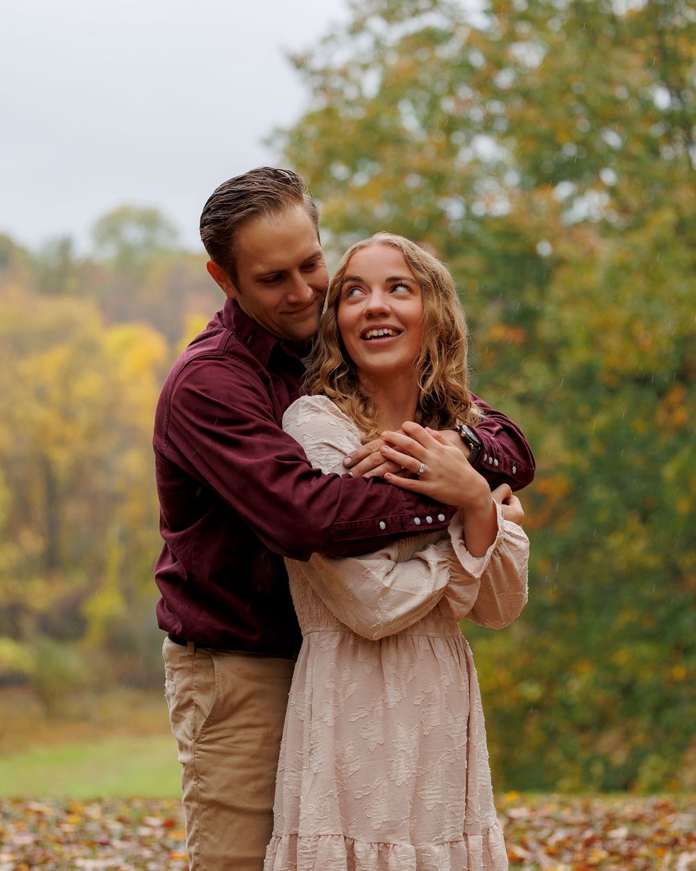 A couple in autumn attire shares a playful moment among fallen leaves in a park setting.