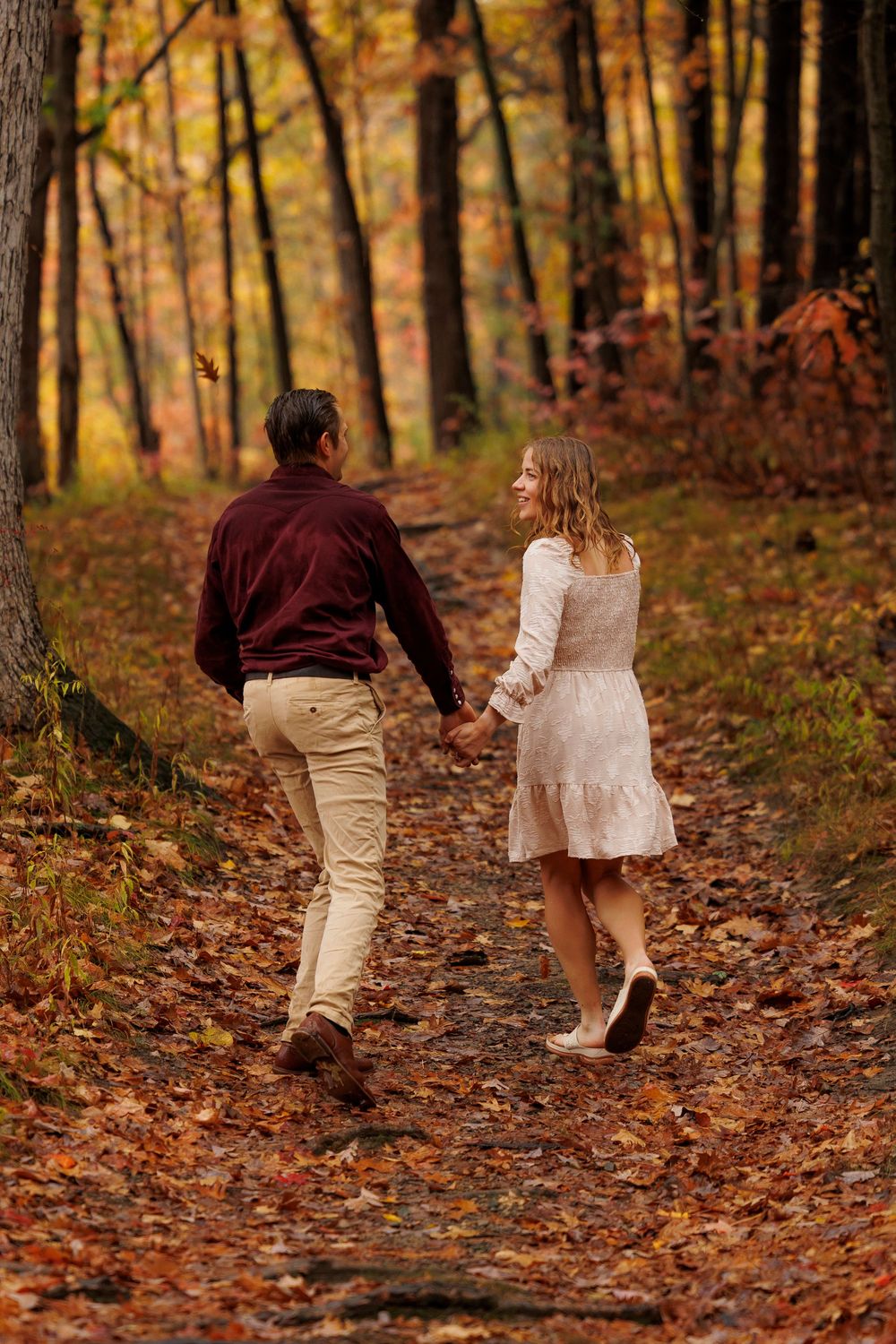 A couple holding hands while walking down a leaf-covered forest path in autumn.