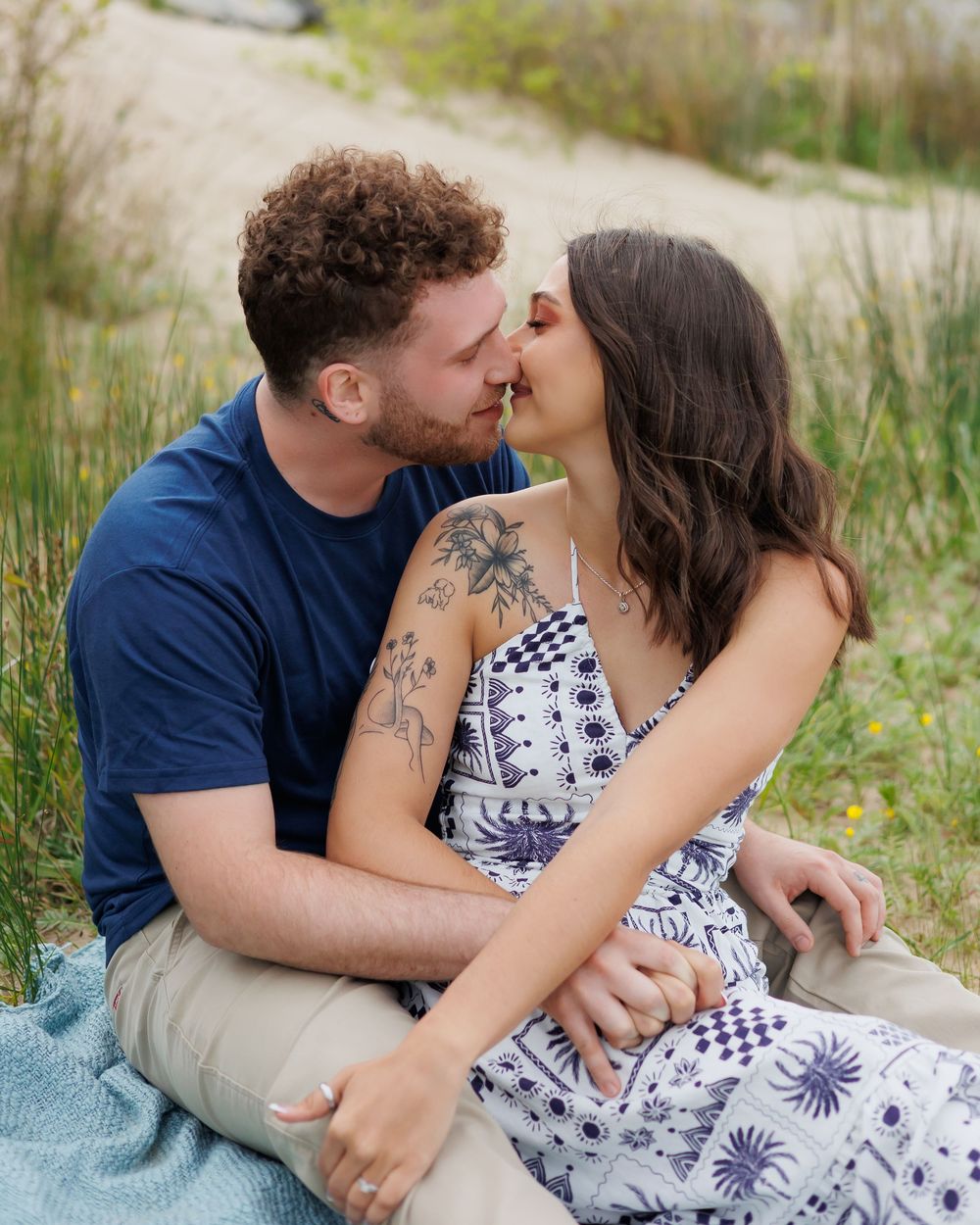 A couple shares an intimate moment while sitting on a blanket outdoors among tall grass on a sunny day.