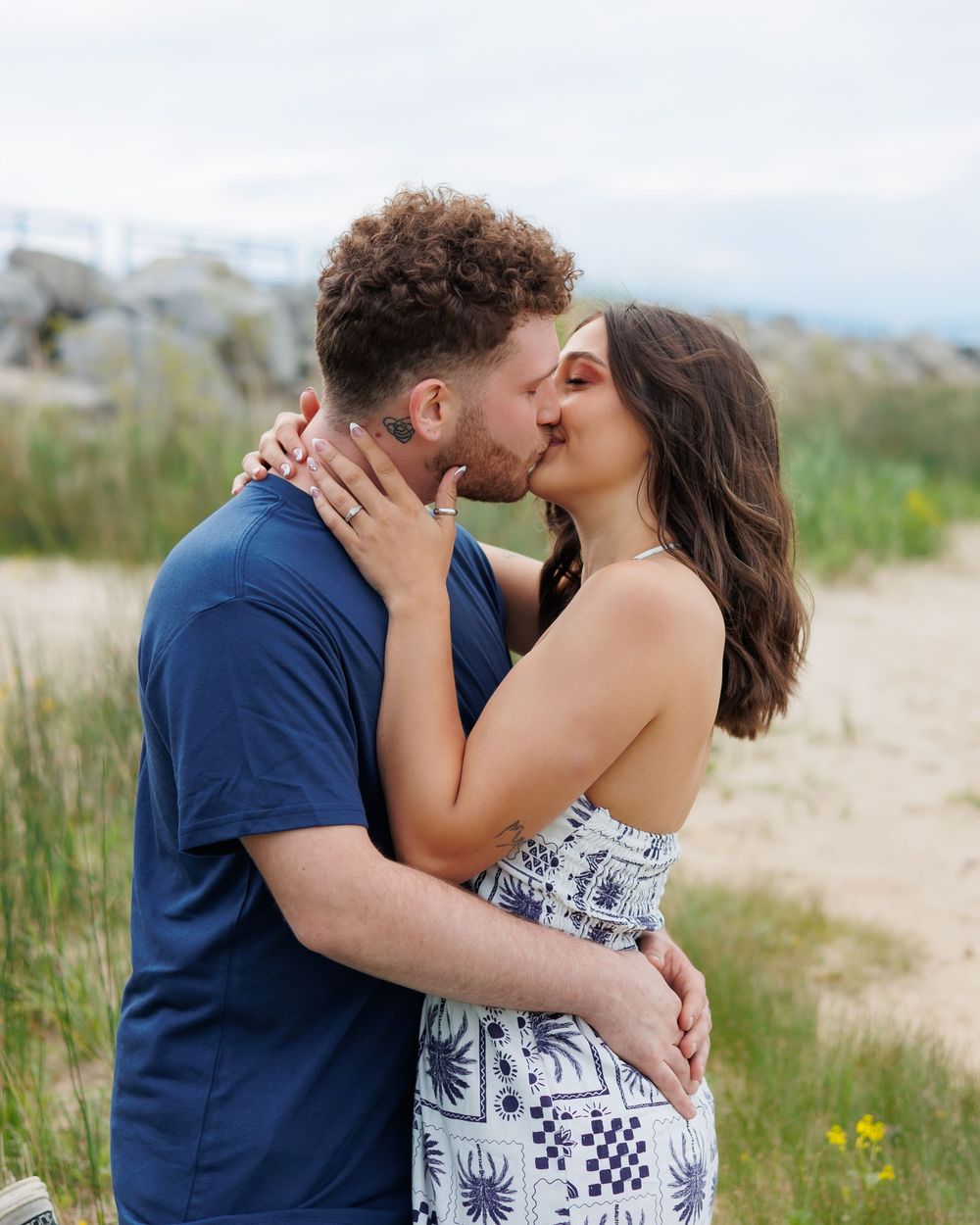 A couple shares a romantic kiss while embracing outdoors with tall grass and mountains in the background.