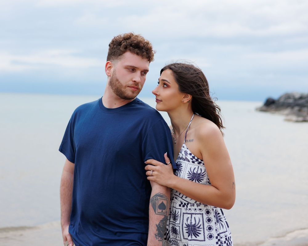 A couple in casual summer attire embraces on a misty beach with rocky coastline in the background.