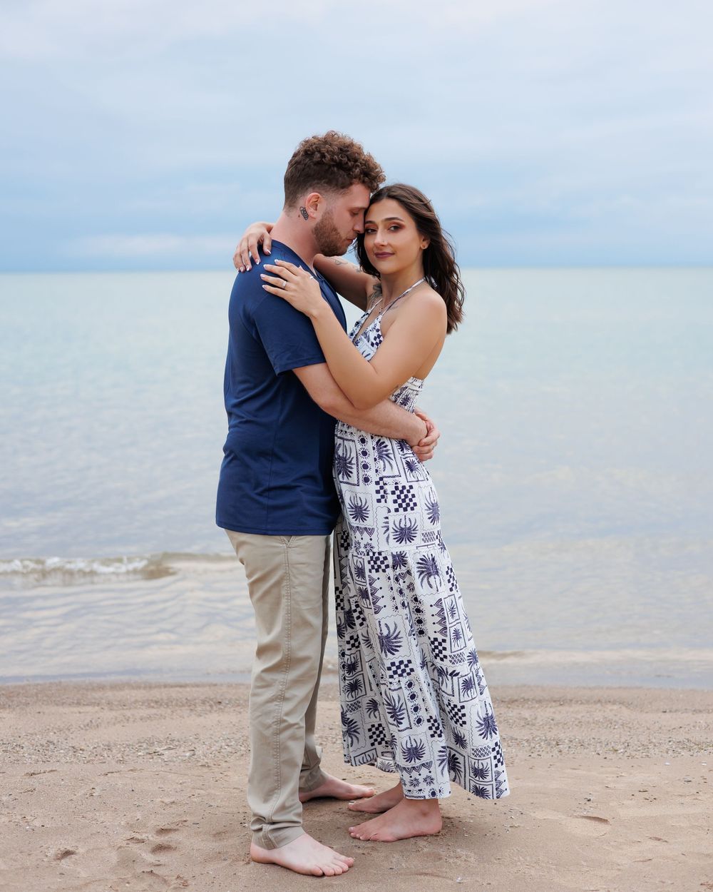 A romantic couple embraces on a peaceful beach with gentle waves and cloudy sky in the background.