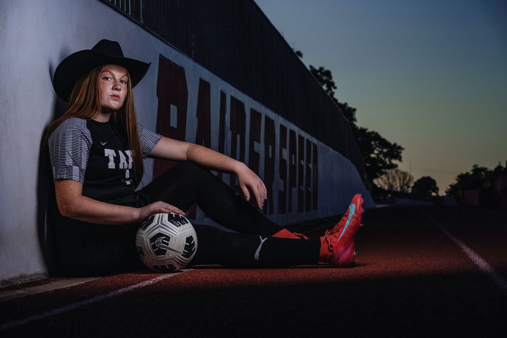 Soccer player in black hat and dark t-shirt sits against wall with ball and pink cleats at sunset.