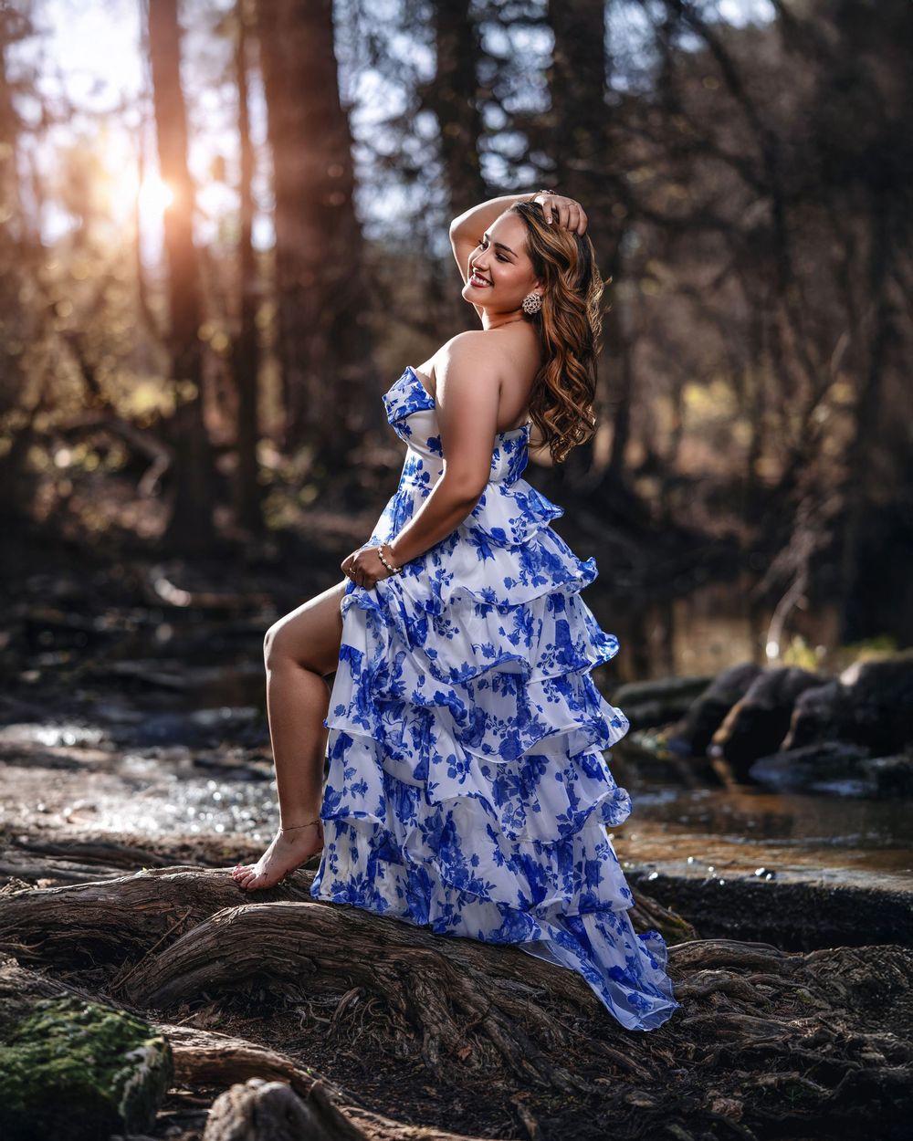 Woman in blue floral ruffled gown poses on rocks in sunlit forest setting.