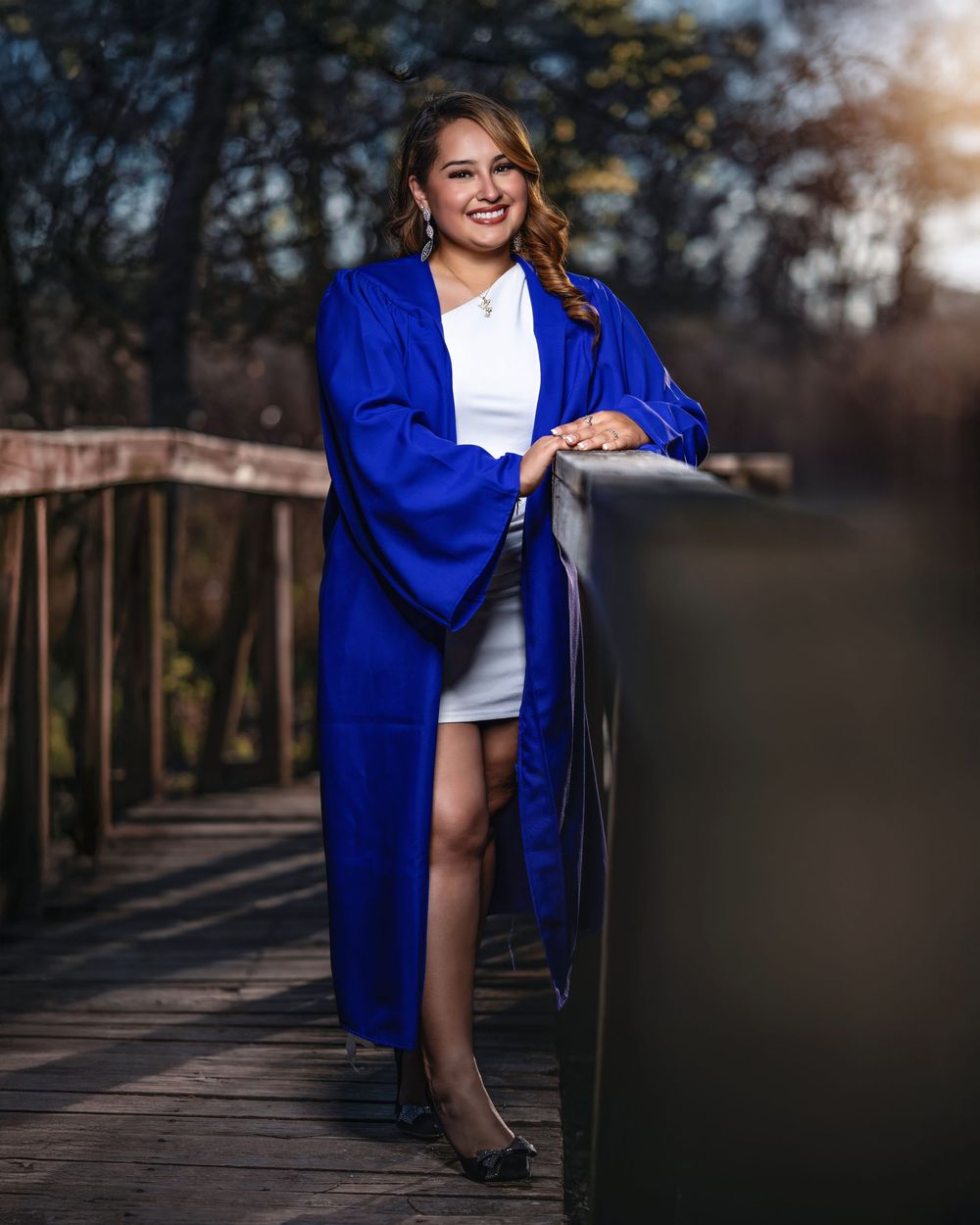 Young woman in blue graduation gown smiling on a wooden bridge, celebrating academic achievement outdoors.