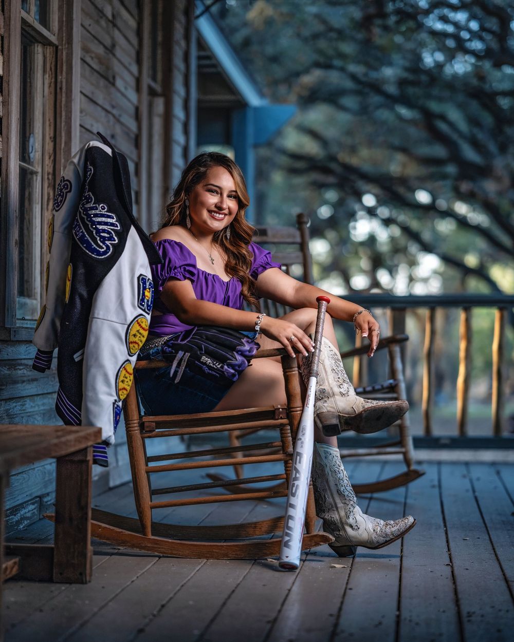 Young woman in purple top and cowboy boots sits smiling in rocking chair on rustic porch holding a baseball bat.