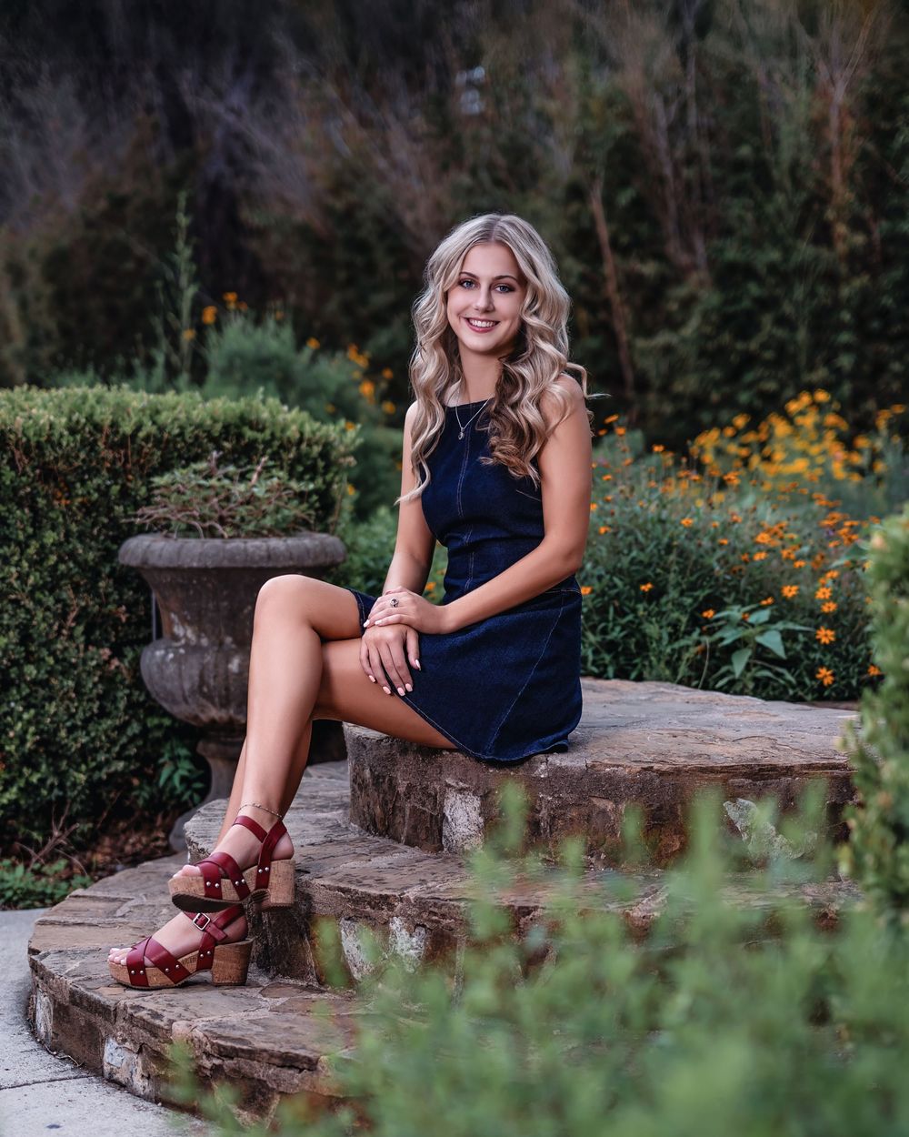 Young woman in navy dress and red heeled sandals sitting on stone steps surrounded by lush garden greenery and yellow flowers.