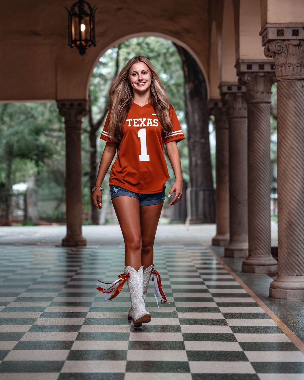 Young woman in Texas Longhorns jersey and white cowboy boots walks confidently through an arched stone colonnade.