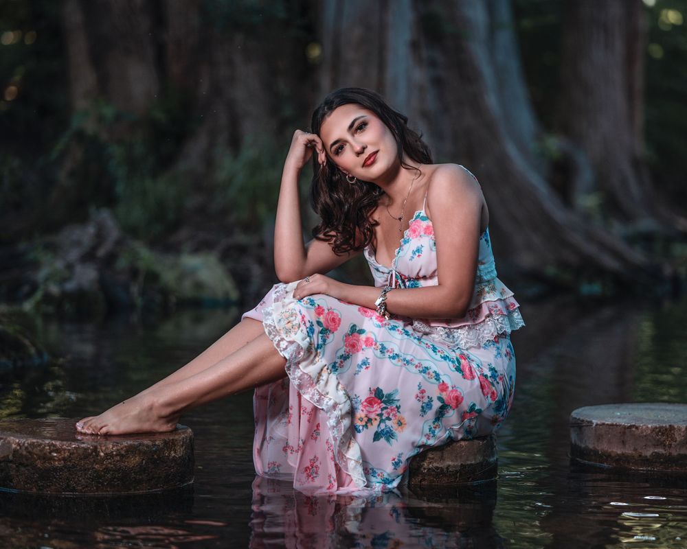 Woman in floral dress sitting on rocks in a serene forest pool, posing gracefully with bare feet in water.