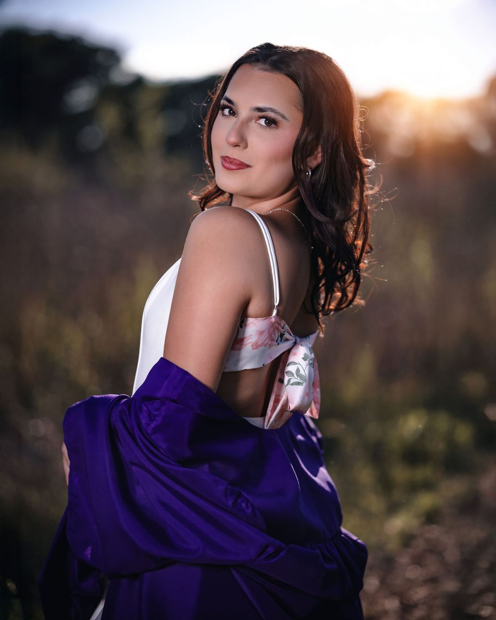 Young woman in white dress and purple satin jacket poses outdoors at golden hour with bokeh background.