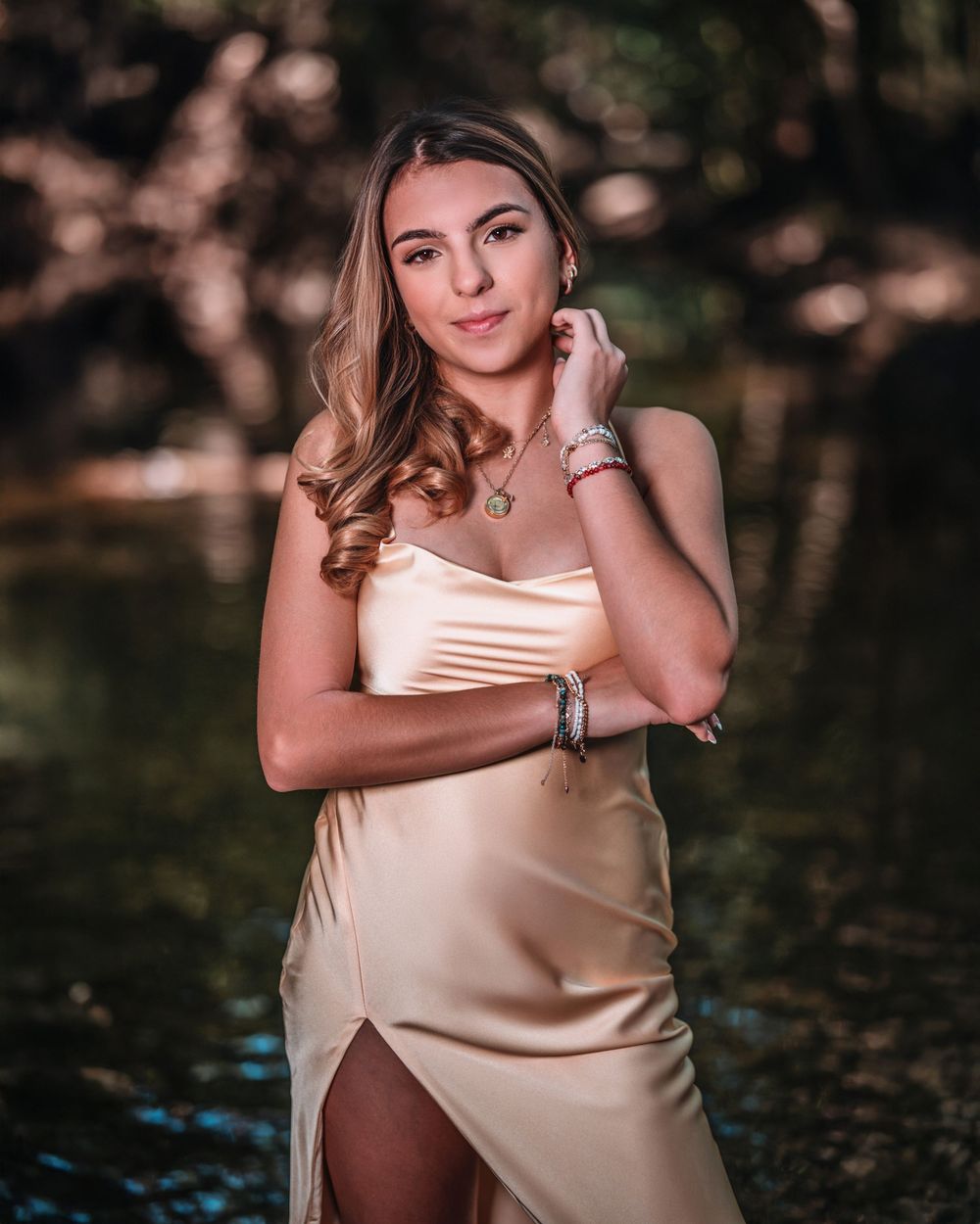 Young woman in elegant gold satin gown posing near water in a lush forest setting.