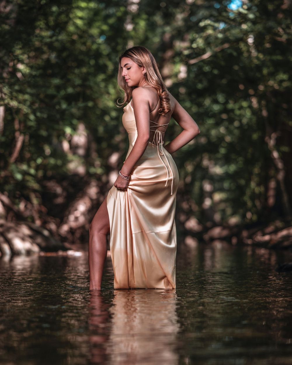 Woman in gold satin gown standing in a forest stream surrounded by lush green trees.