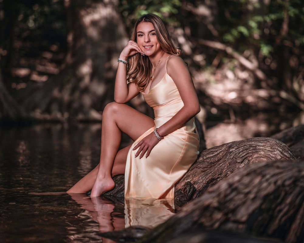 Young woman in gold dress sitting on log in forest stream, smiling.