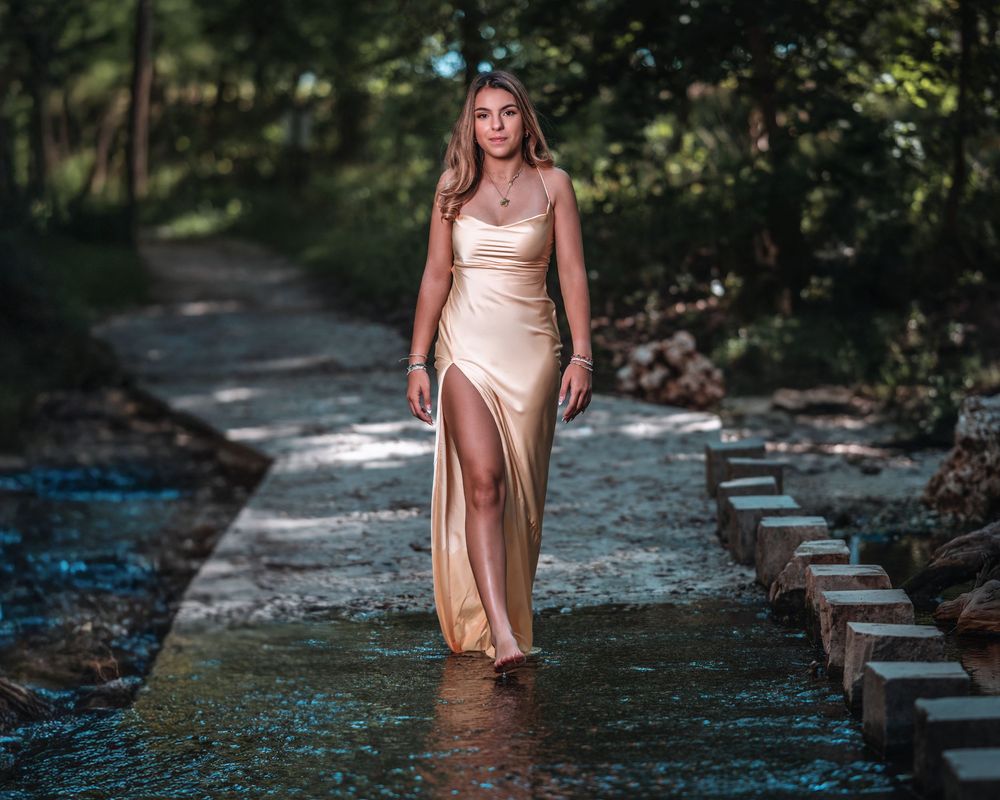 Young woman in elegant gold satin gown walking through shallow stream on stepping stones surrounded by lush greenery.
