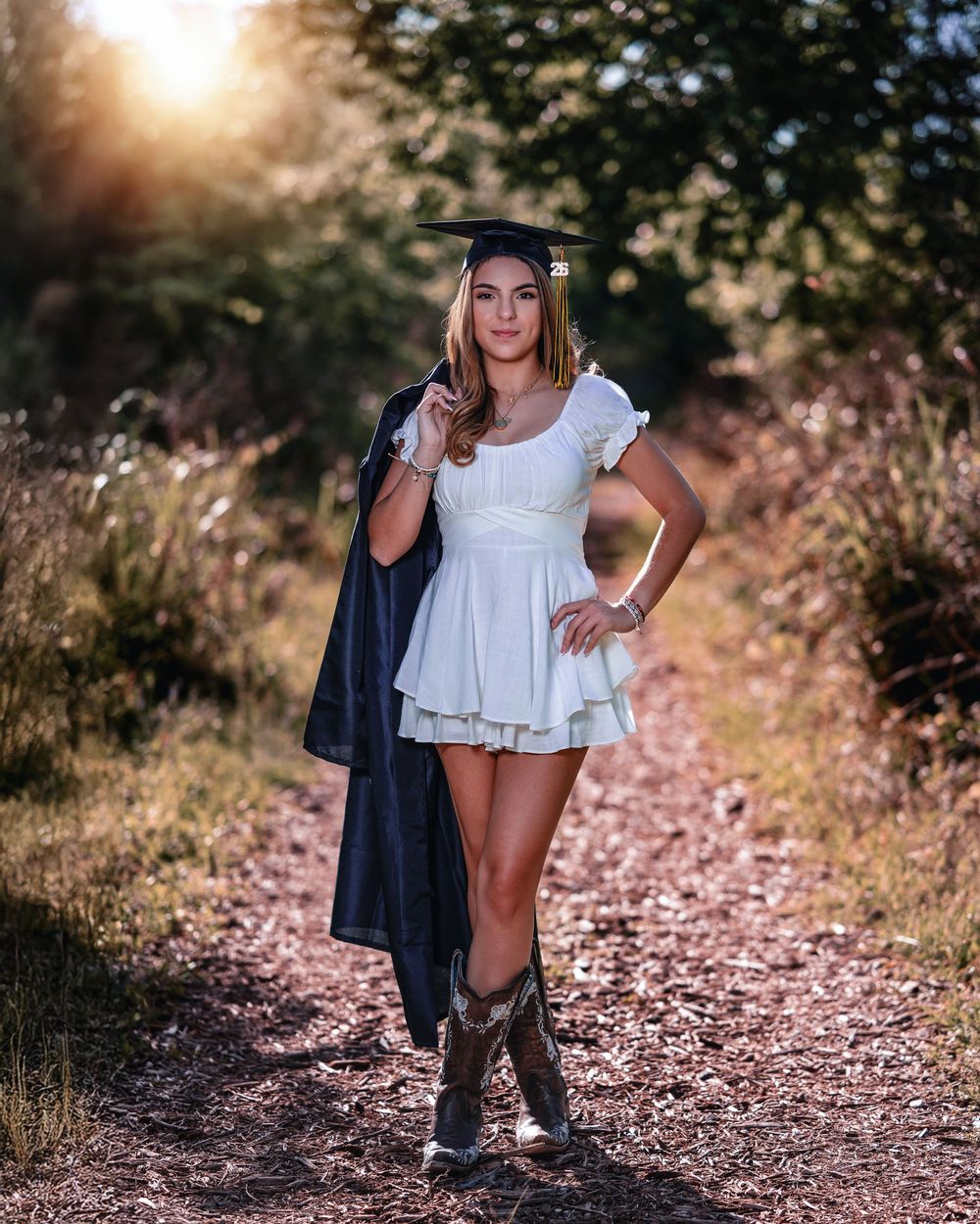 Graduate in white dress and cowboy boots holding cap and gown on sunlit forest path.