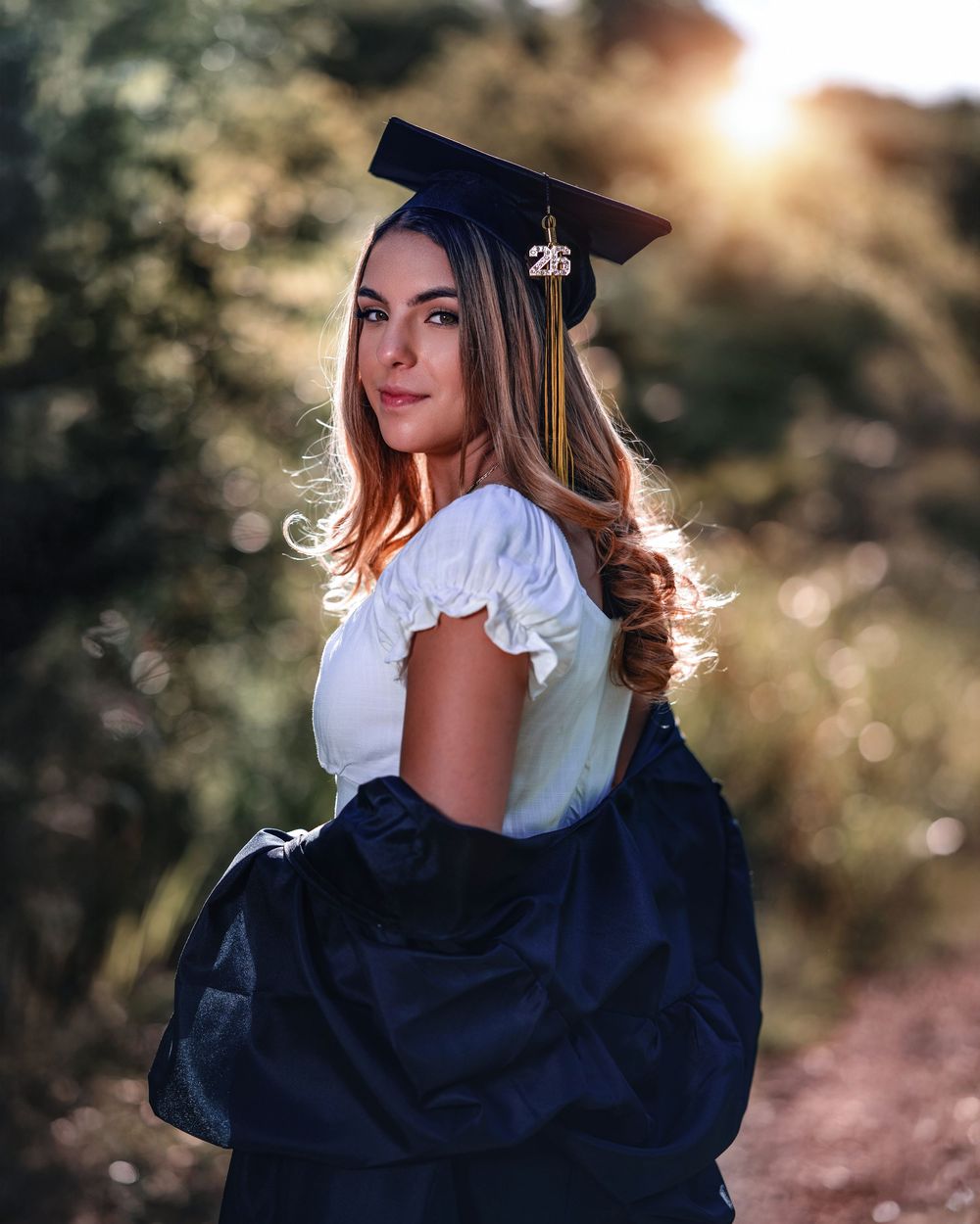 Young woman in graduation cap and gown poses outdoors in golden sunlight, smiling over her shoulder.