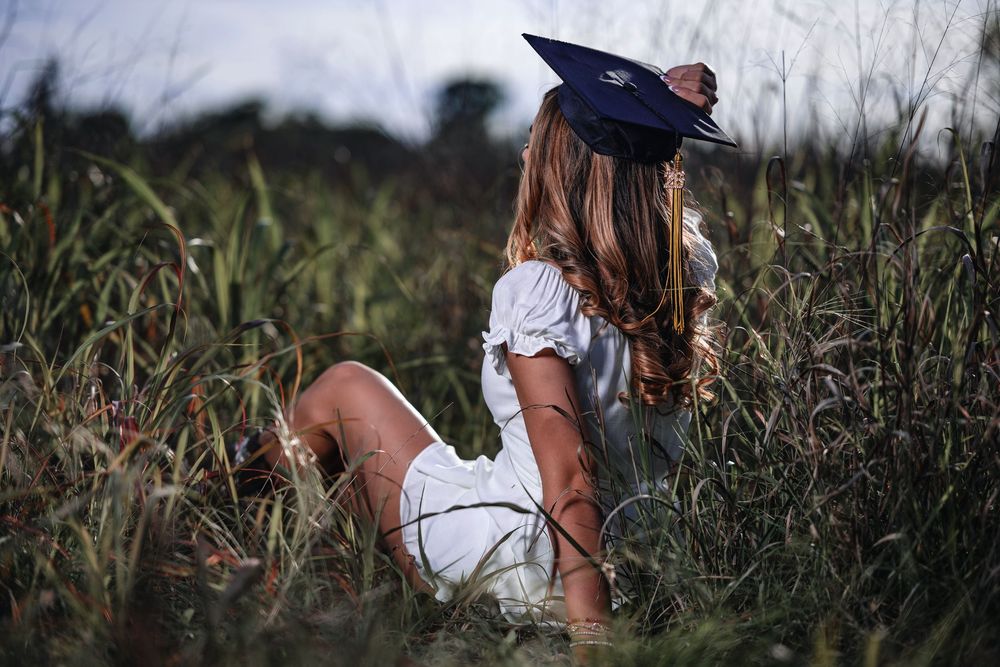 Graduate in white dress and cap sitting in tall grass field at dusk.
