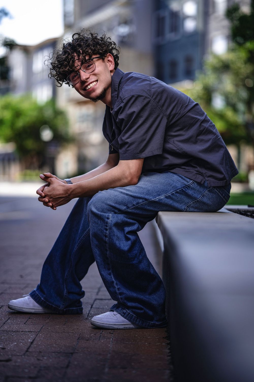 Young individual with curly hair and glasses wearing a navy shirt and jeans sits casually on a concrete ledge outdoors.