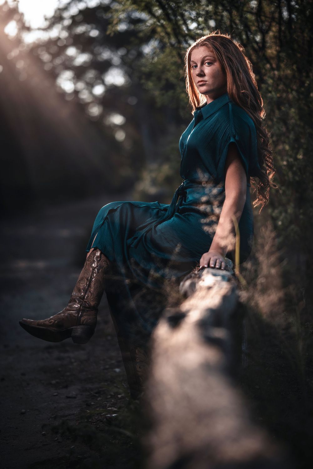 A figure in a teal vintage dress poses on a rustic log at sunset wearing brown cowboy boots in a moody forest setting.