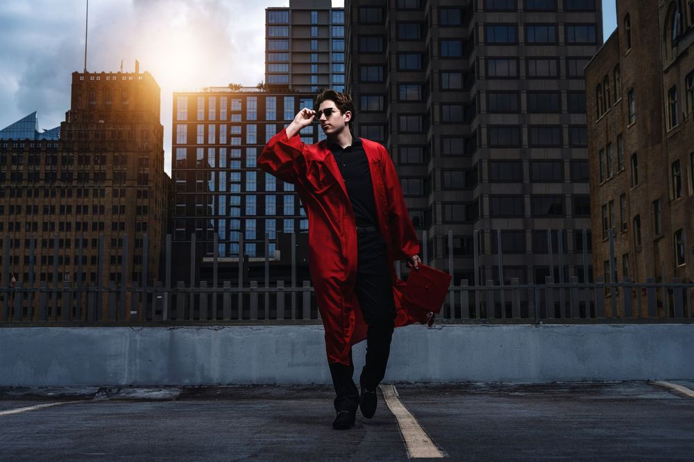 Person in bright red coat walking dramatically on urban rooftop against sunset and city buildings.