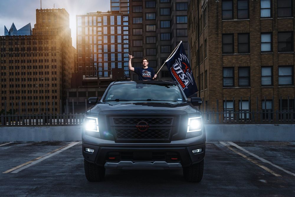 A black pickup truck with headlights on stands in an urban parking lot at sunset with skyscrapers in the background.