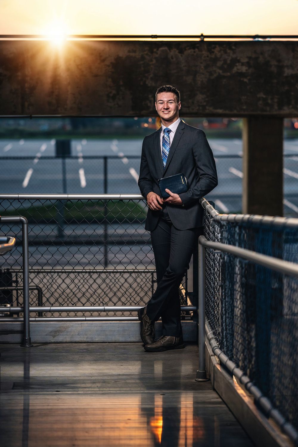 Professional in a dark suit and tie stands on a walkway at sunset with an empty parking lot in the background.