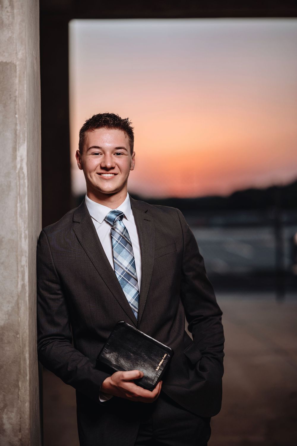 Professional portrait of someone in a dark suit and plaid tie holding a book at sunset against a blurred outdoor background.
