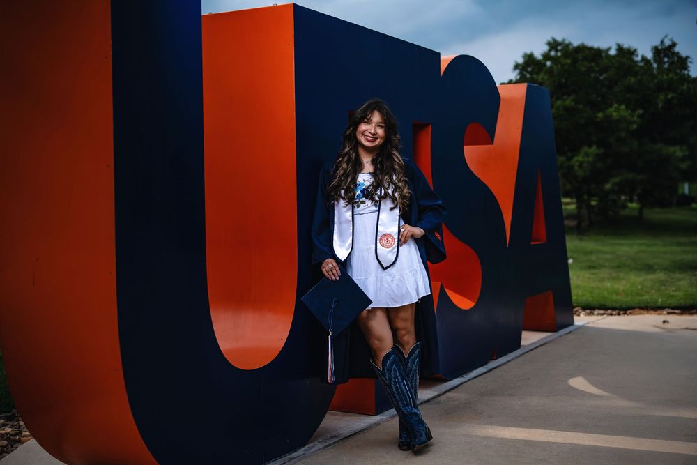 A student in a dark jacket and cowboy boots poses in front of large colorful USA letters on a college campus.