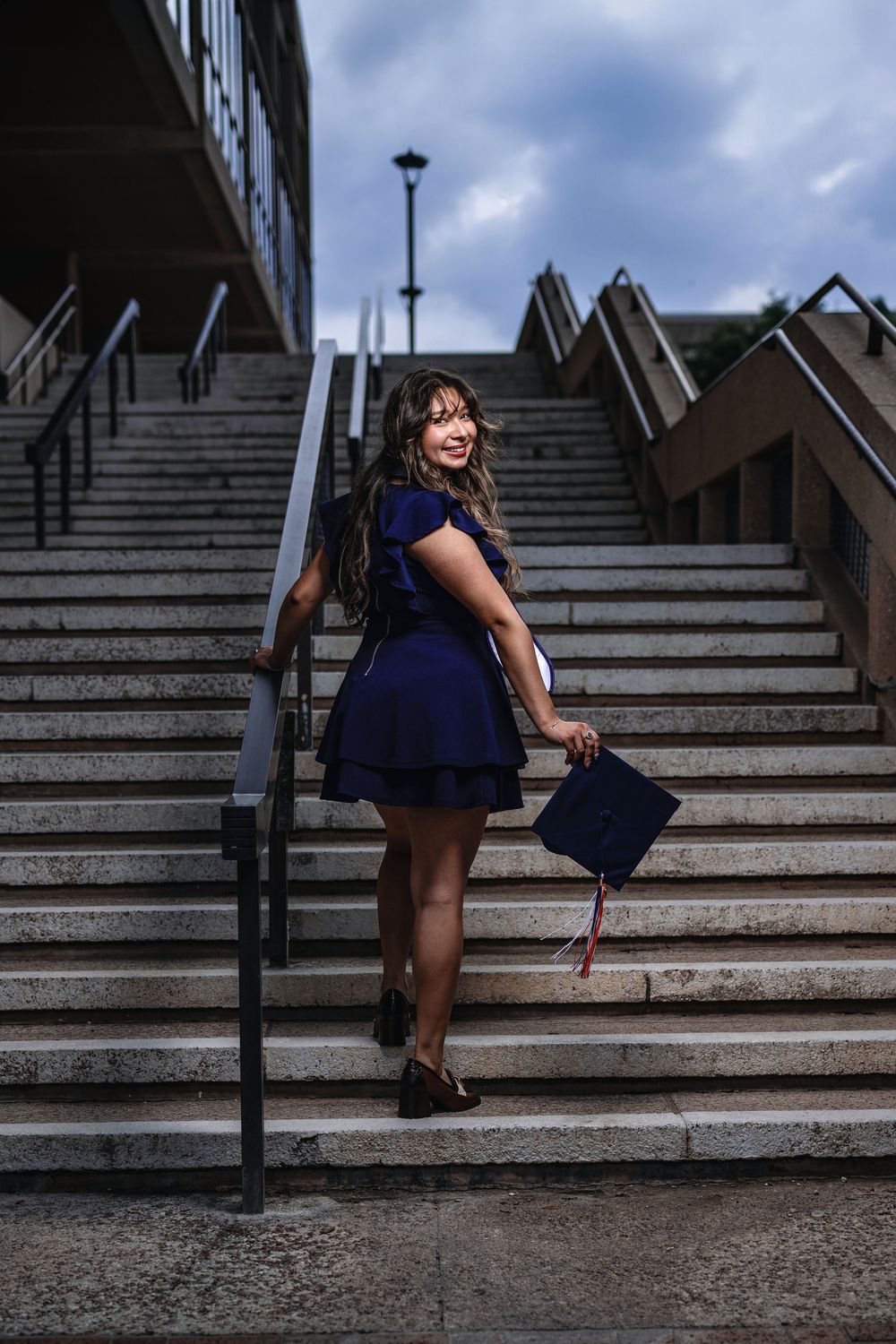 Graduate in navy blue dress smiles while standing on concrete steps holding graduation cap and tassel.