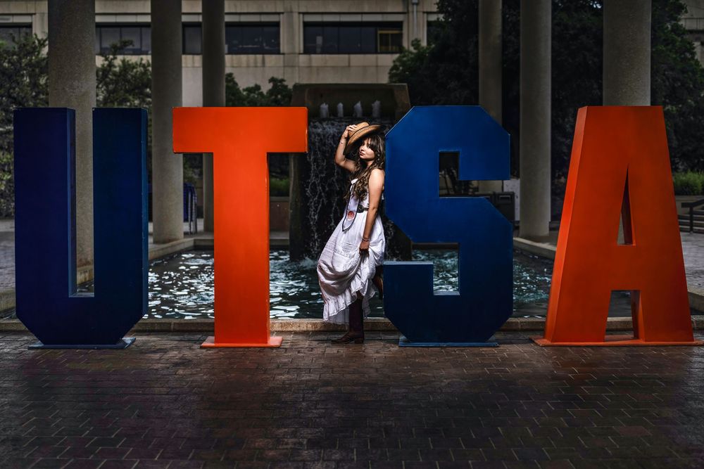 Large colorful UTSA letters serve as backdrop for elegant figure in white dress against urban building setting.
