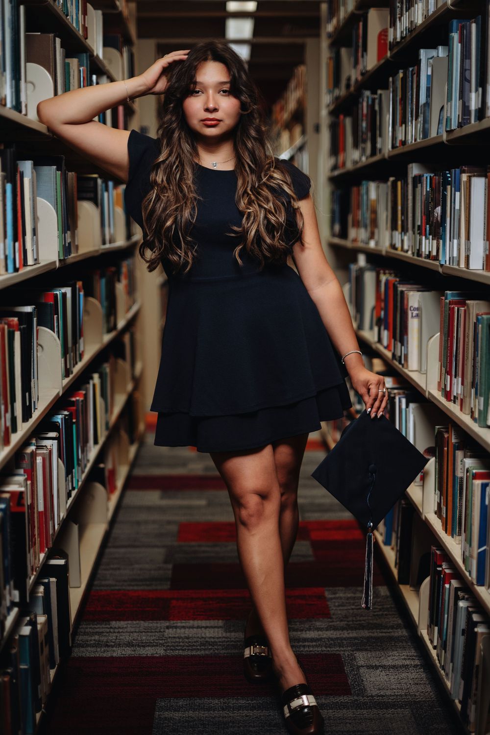 Person in a black dress posing between library bookshelves with carpeted floors and soft lighting.
