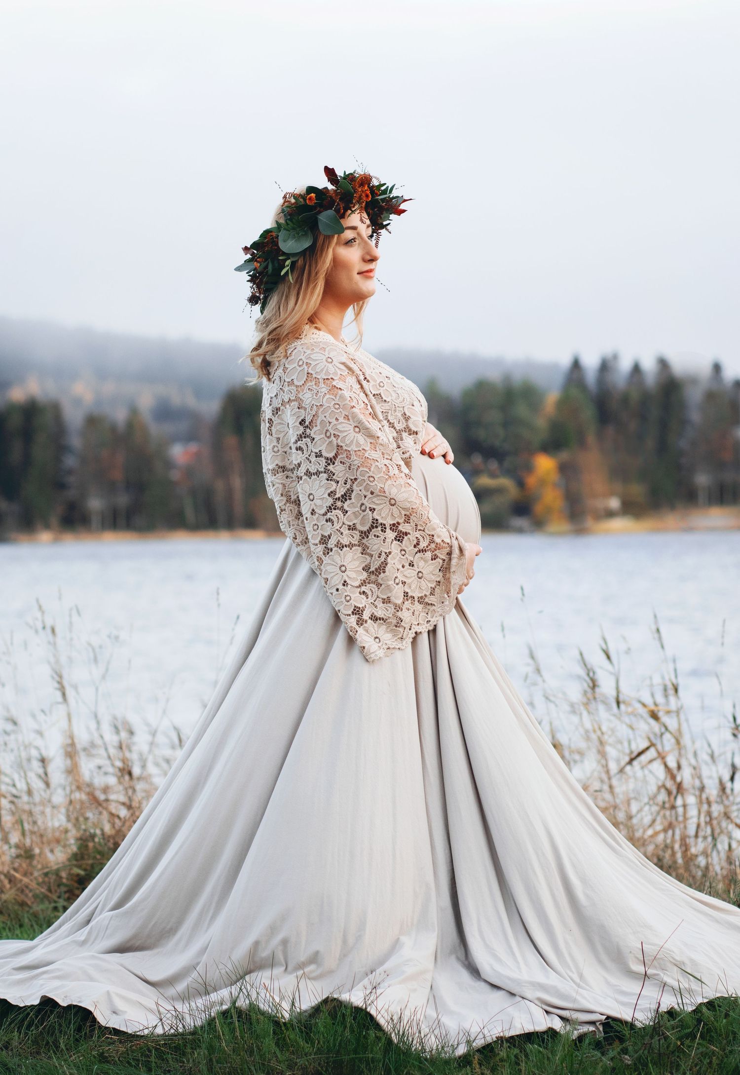 Pregnant woman in lace top and gray skirt with autumn floral crown by lake.