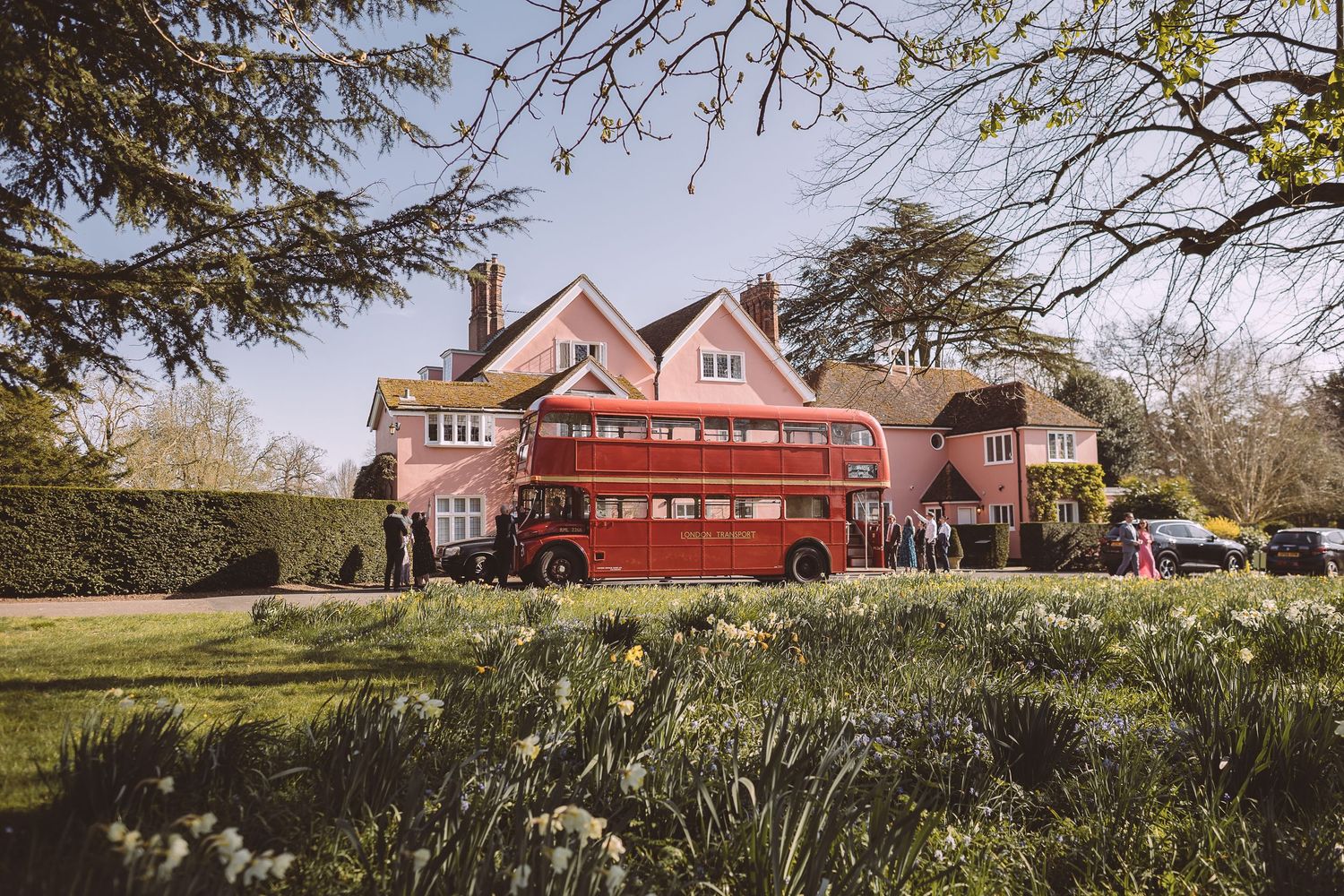 A red and pink country house stands behind a spring garden with blooming daffodils and surrounding trees. A routemaster stands outside.