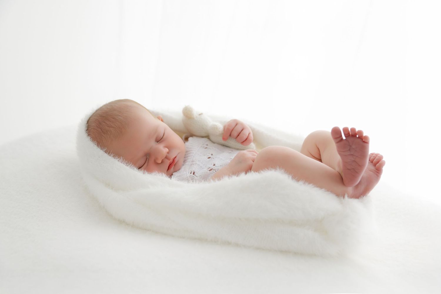Baby wearing white onesie laying on white bed sheets while stretching and moving.