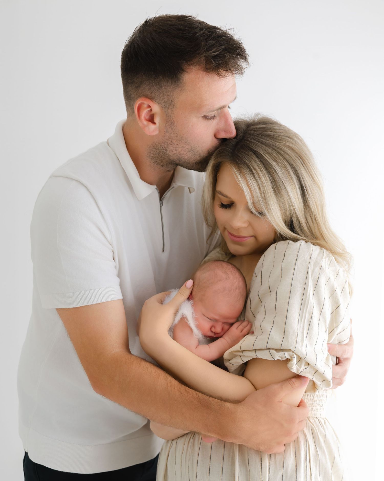 A tender family moment shows parents embracing their newborn baby in a bright studio setting.