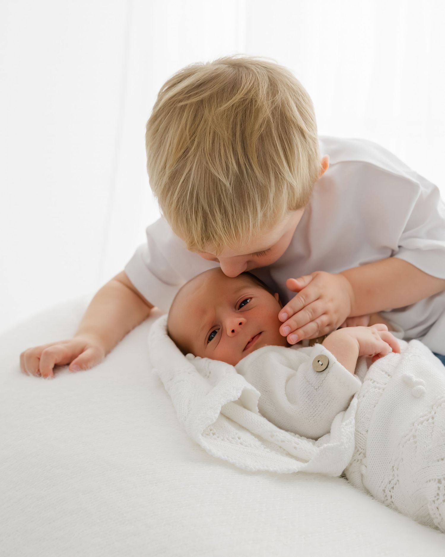 A young child in white lovingly kisses a newborn baby sibling wrapped in a soft white blanket.