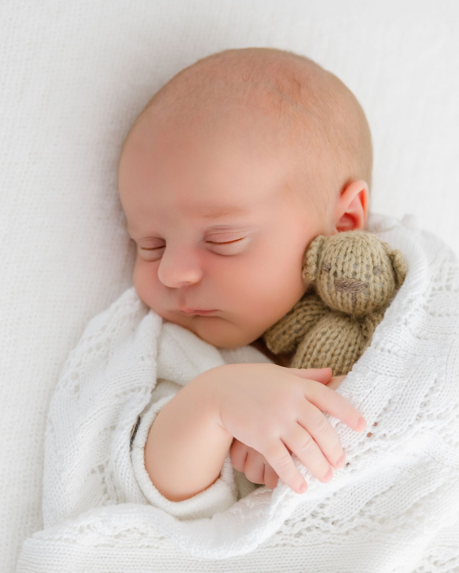 A sleeping newborn baby wrapped in soft white lace blanket snuggles against a knit teddy bear.