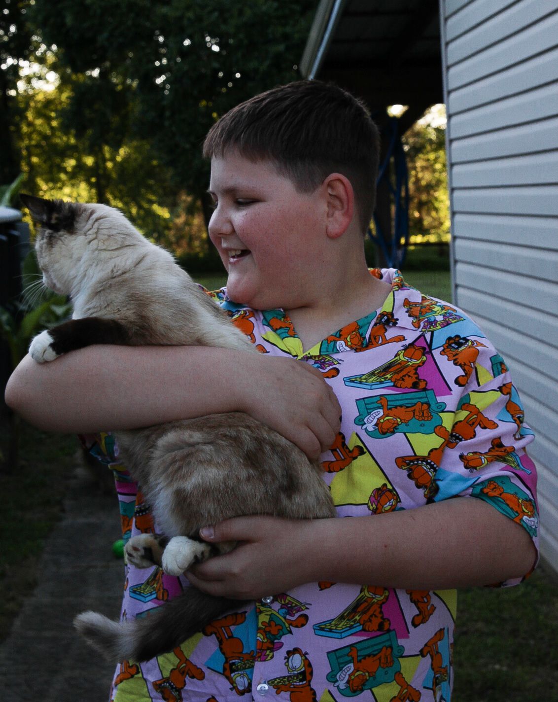 Young person in colorful patterned shirt holds and hugs a fluffy white and grey cat outside near a house wall.
