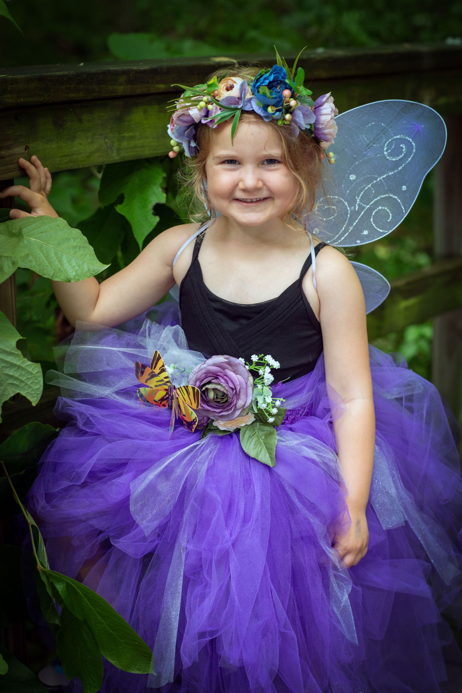 A child in a fairy costume wearing purple tulle tutu, floral crown and translucent wings poses in a garden setting.