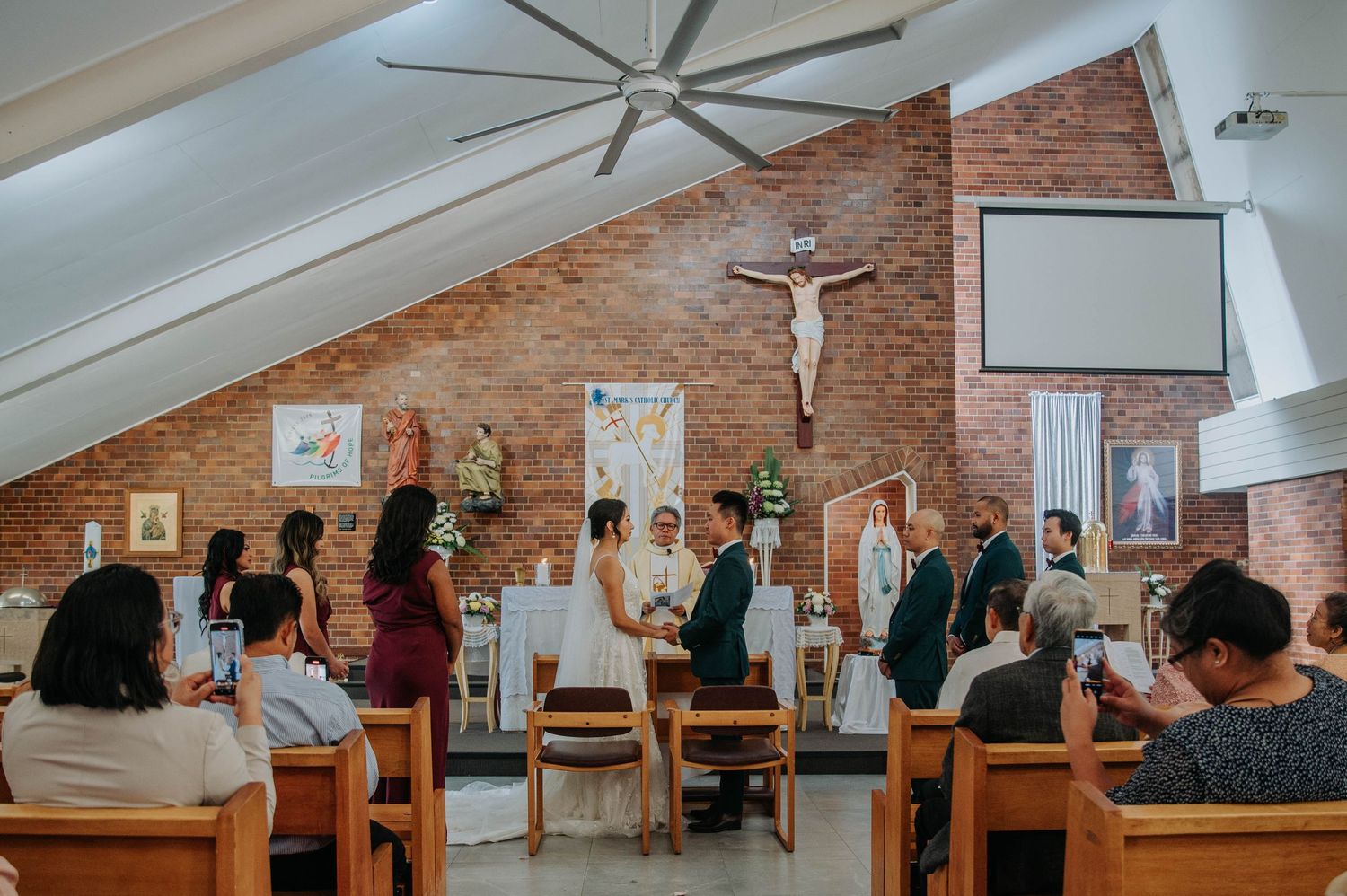 A group of people seated in wooden pews inside a church with exposed brick walls and a large crucifix mounted in front.