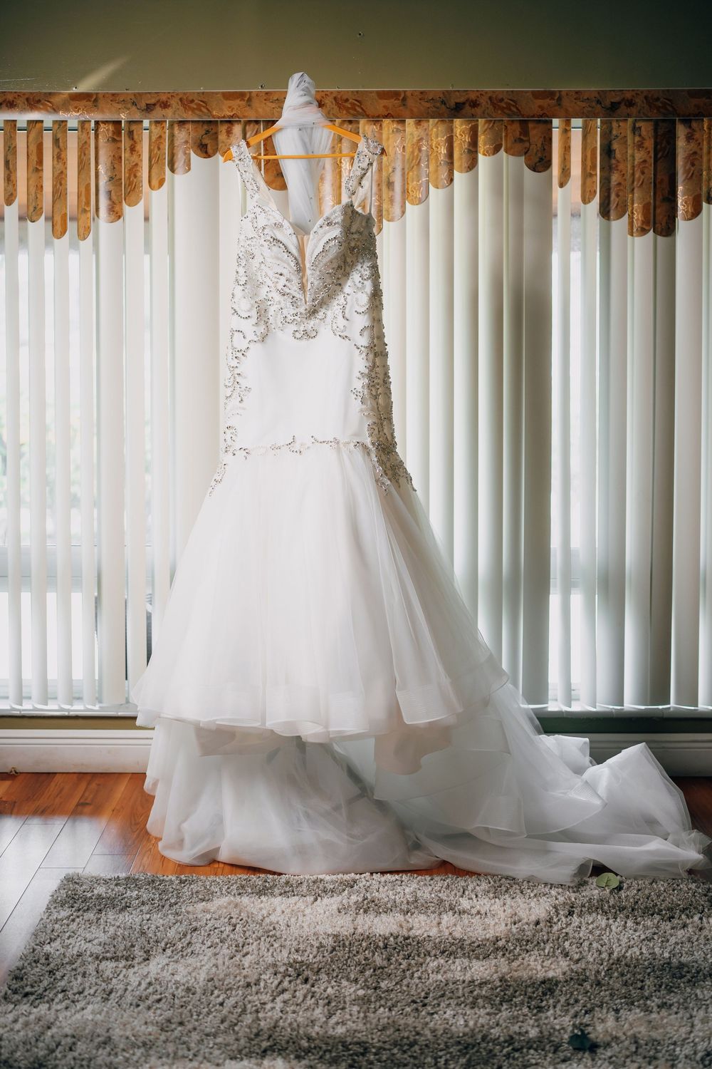 Elegant white lace wedding dress with beaded bodice and tulle skirt displayed against vertical blinds.